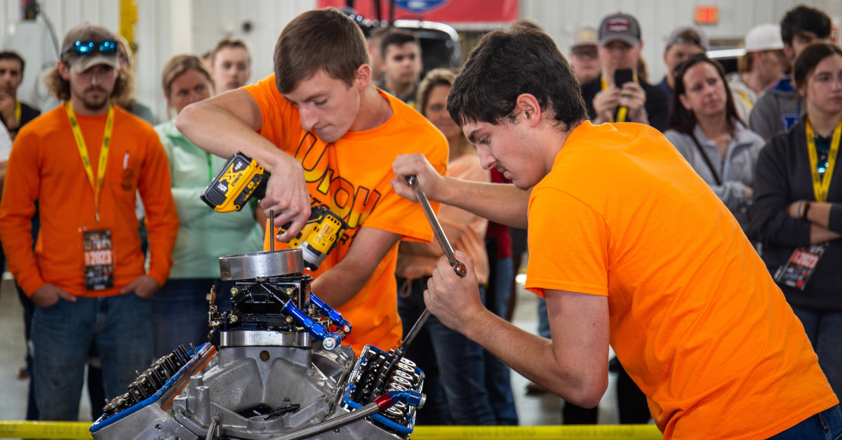 A crowd gathers around a group of UNOH students working on an engine